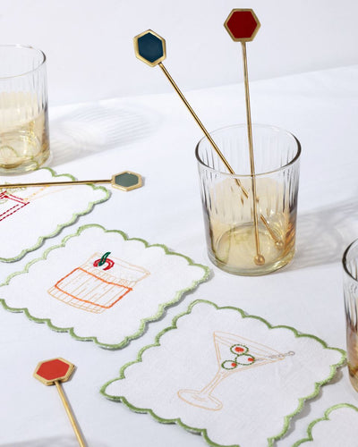 Decorative cocktail stirrers with hexagonal ends on a glass of drink, surrounded by embroidered coasters.